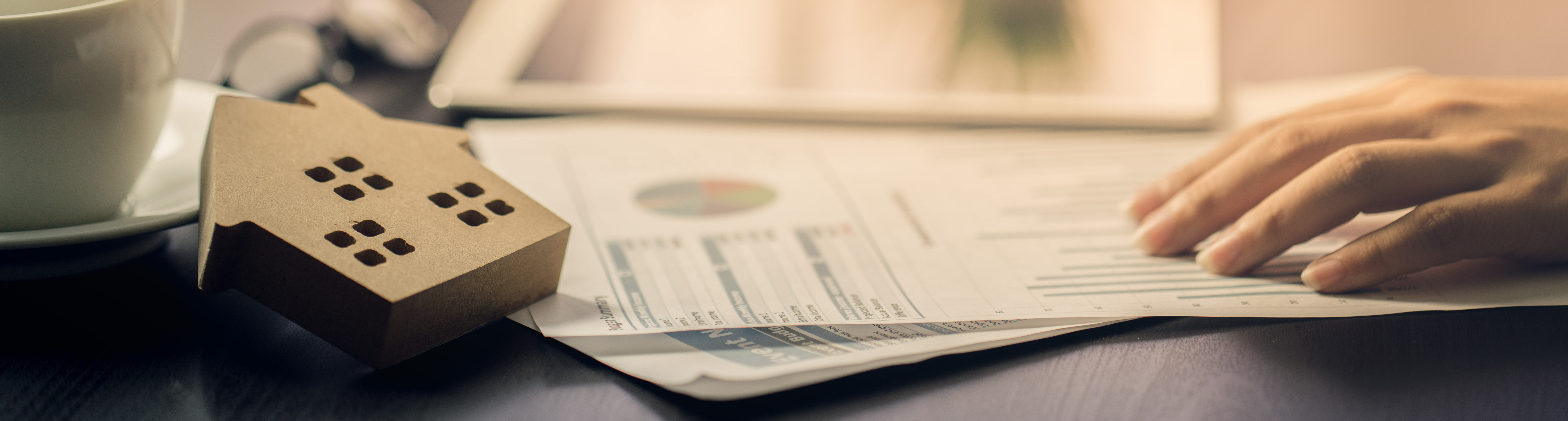Hand holding report papers with small wooden cut-out of house on desk.